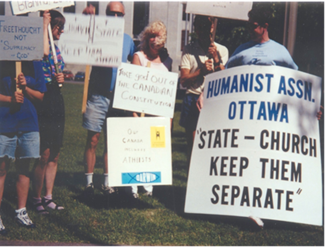 A small, dedicated group of secular demonstrators stands together, holding hand-painted, black-and-white signs and placards advocating for the separation of church and state.